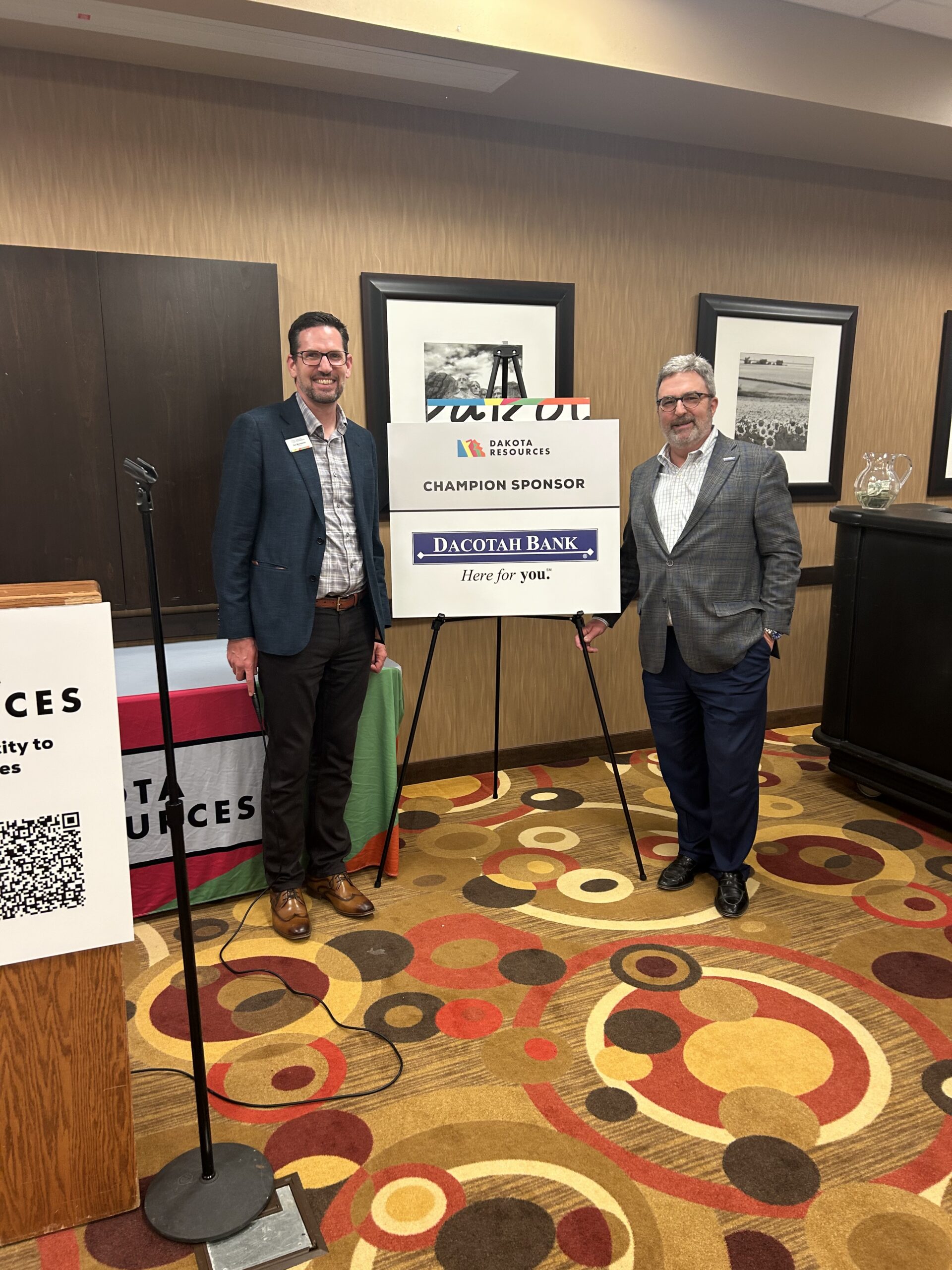 Two men stand indoors on either side of a sponsor sign on an easel at a formal event. The sign reads “Dakota Resources – Champion Sponsor” with the Dacotah Bank logo and the tagline “Here for you.” Both men smile toward the camera and wear business attire. A podium with a microphone, framed wall art, and patterned carpet are visible in the conference-room setting.