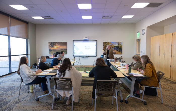 Facilitator leading a small group workshop in a conference room, with participants seated around tables and a presentation displayed on a screen.