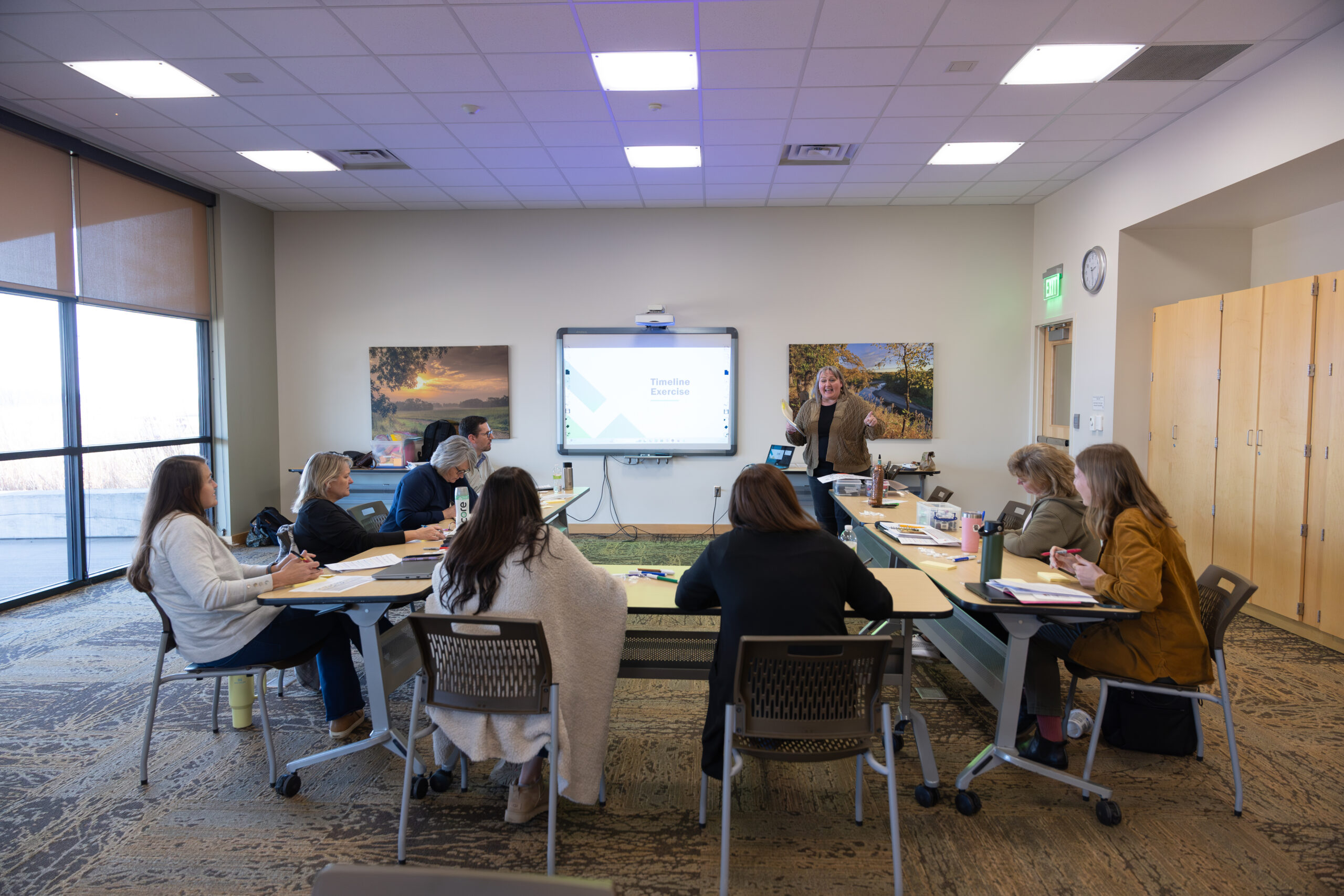 Facilitator leading a small group workshop in a conference room, with participants seated around tables and a presentation displayed on a screen.